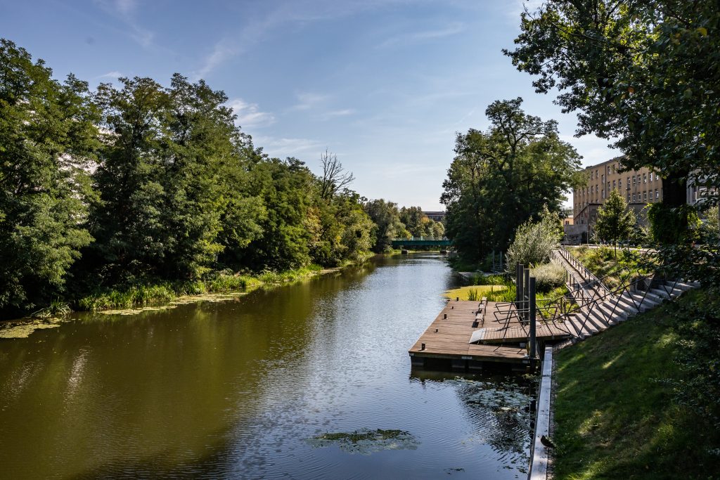 Promenada nad Oławą - Widok na rzekę i most, po prawej stronie drewniana kładka z trzema ławkami i schody, po lewej stronie towarzyszące zadrzewienie.