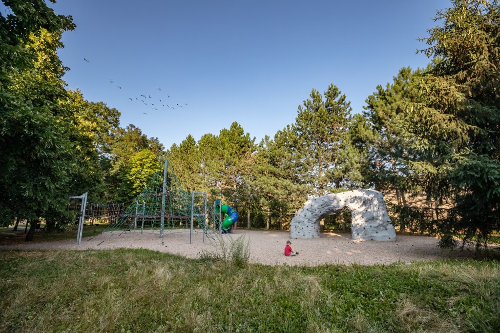 Park przy ul. Strachocińskiej- widok na plac zabaw: liny i ścianka wspinaczkowa oraz zakręcona zjeżdżalnia, dookoła zadrzewienie.