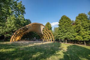 Park przy ul. Strachocińskiej- widok na drewnianą scenę, obok okazy drzew, na drugim planie fragment placu zabaw.