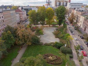 Skwer Sybiraków - widok z lotu ptaka na skwer, na pierwszym planie zieleń: okazy drzew i krzewów. Na drugim planie betonowy plac ze stołami do gry w szachy.