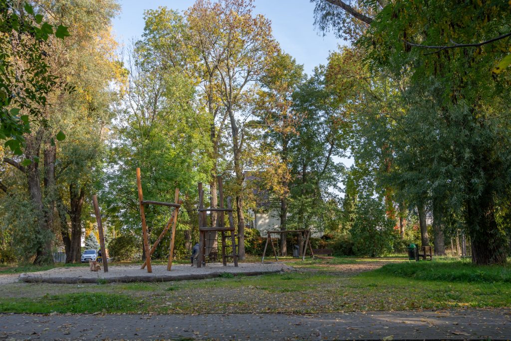 Skwer przy ul. Rubinowej-Nefrytowej- widok na plac zabaw, na pierwszym planie drewniane sprzęty do wspinaczki, na drugim planie huśtawka i towarzyszące zadrzewienie.