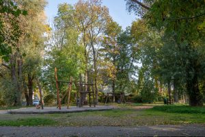 Skwer przy ul. Rubinowej-Nefrytowej- widok na plac zabaw, na pierwszym planie drewniane sprzęty do wspinaczki, na drugim planie huśtawka i towarzyszące zadrzewienie.