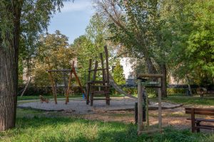 Skwer przy ul. Rubinowej-Nefrytowej- zbliżone ujęcie na plac zabaw, widoczne sprzęty do wspinaczki, metalowa zjeżdżalnia i bujaki, w tle zadrzewienie.