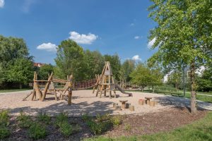 Park Wojszyce- widok na drewniany plac zabaw, widoczne dwa małe stoliki z krzesłami, wieża ze zjeżdżalnią i elementy do wspinania.