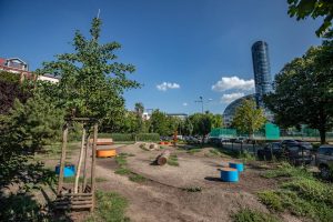 Skwer przy ul. Grabskiego- na pierwszym planie młode drzewo, na drugim planie widoczny tunel i pień do zabaw, niewielkie wzniesienia oraz okrągłe siedziska, w oddali Sky Tower.