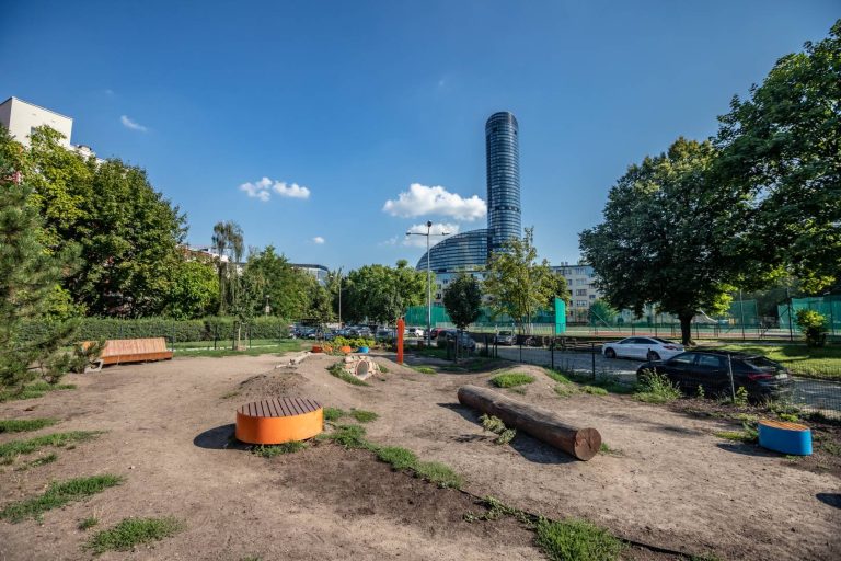 Skwer przy ul. Grabskiego- zbliżenie na małą architekturę (okrągłe siedziska, tunel, pień), na drugim planie zieleń towarzysząca, w oddali Sky Tower.