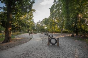 Park Tadeusza Różewicza - widoczny park dla psów, na pierwszym planie ścieżka oraz przeszkody wykonane z opon przymocowanych do drewnianych słupów, na drugim planie inne sprzęty do zabaw.
