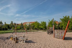 Park Migdałowy- widok na plac zabaw, widoczna drewniana platforma do balansowania, huśtawki i ścianka wspinaczkowa.