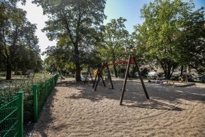 Park generała Mariana Langiewicza- widok na plac zabaw: drewniana huśtawka, piaskownica, bujak, na drugim planie zadrzewienie.