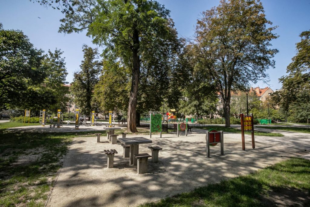 Park generała Mariana Langiewicza- widok na stoły do gry w szachy, na drugim planie siłownia oraz plac zabaw.