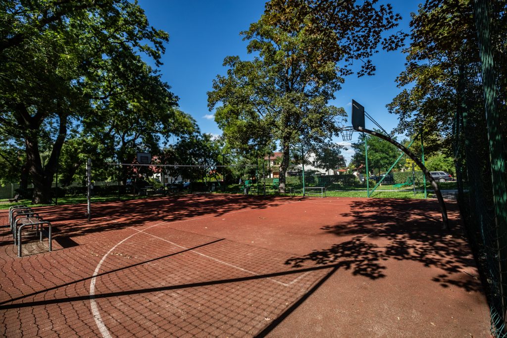 Skwer przy ul. Stanisławowskiej- ujęcie na wnętrze boiska sportowego, widoczne kosze do gry w koszykówkę, ławki i metalowe ogrodzenie.