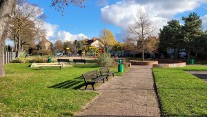 Skwer przy ul. Stanów Zjednoczonych- widok na okrągły plac, pośrodku okaz drzewa, po lewej stronie ławki. W tle widoczny plac zabaw i zadrzewienie.