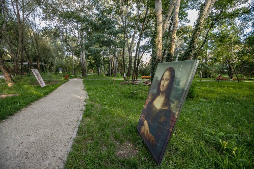Park Leonarda da Vinci- na pierwszym planie widoczna ścieżka i obraz Mona Lisa, na drugim planie autoportret Leonarda da Vinci i zadrzewienie.