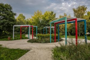 Park Leonarda da Vinci- widok na kolorowe pergole, pośrodku rabata bylinowa, w tle zadrzewienie.