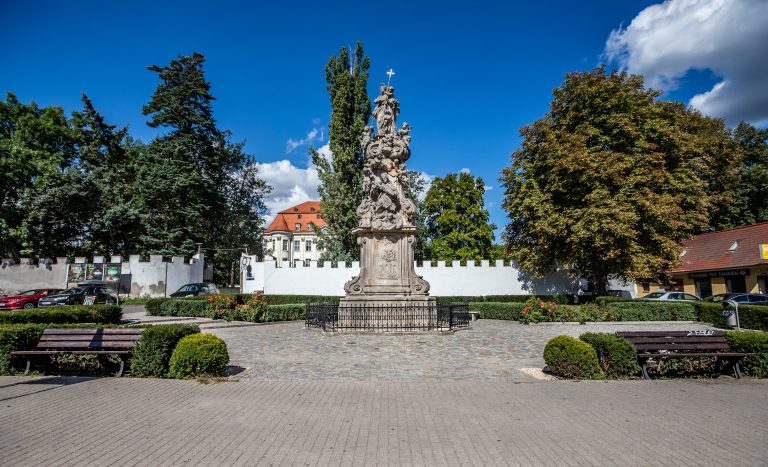 Skwer przy pl. Świętojańskim- widok na rzeźbę Matki Boskiej z Dzieciątkiem i Świętego Jana Nepomucena i otaczające ją żywopłoty, na drugim planie widoczne wejście do Parku Leśnickiego oraz fragment zamku.