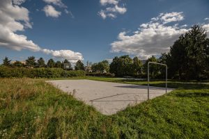 Skwer przy ul. Śnieżnej- widok na nieogrodzone boisko sportowe. Na pierwszym planie polana, kosz do gry w koszykówkę i metalowa bramka. Na drugim planie grupa krzewów, szpaler drzew i stół do gry w tenisa.