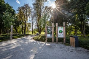 Ekopark na Stabłowicach- widok na wejście do parku. Na pierwszym planie drewniany kosz i dwie tablice informacyjne, na drugim planie sprzęty do zabaw i towarzysząca roślinność.