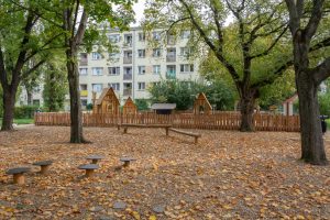 Skwer św. o. Damiana de Veuster - widok na drewnianą małą architekturę, na drugim planie ogrodzony plac zabaw, pojedyncze okazy drzew, w tle blok mieszkalny.