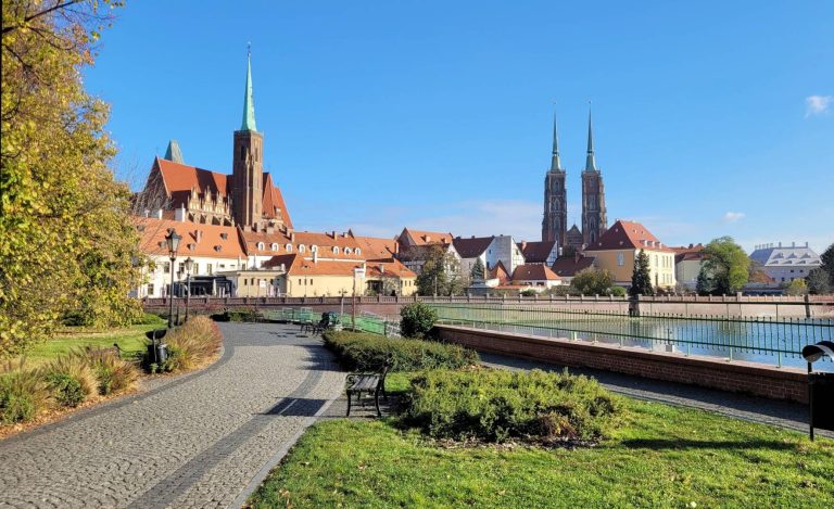 Bulwar Piotra Włostowica - widok na bulwar, po lewej stronie nasadzenia bylin i traw ozdobnych, po prawej stronie grupy krzewów. W tle fragment rzeki oraz Kolegiata pw Świętego Krzyża i św. Bartłomieja.