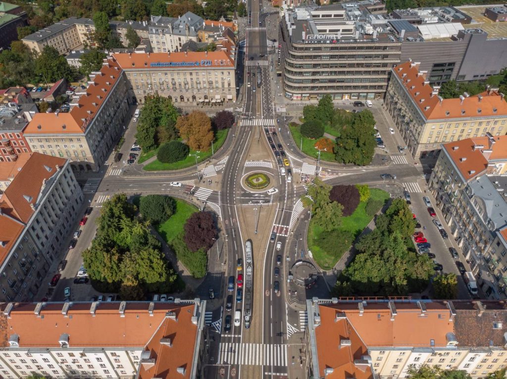 Plac Kościuszki - Widok na ścieżkę kamienną, po obu stronach ogrodzone niskim płotkiem place zieleni z okazami drzew i krzewów.