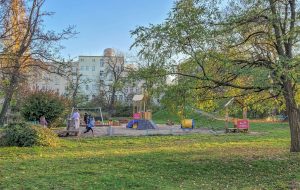 Park Lesława Węgrzynowskiego- ujęcie na plac zabaw, widoczne drewniane domki ze ścianką wspinaczkową, fabryka piasku, huśtawki, na drugim planie pagórek i zadrzewienie.