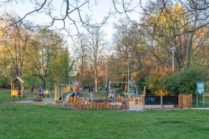 Park Lesława Węgrzynowskiego- zbliżone ujęcie na plac zabaw ogrodzony drewnianymi słupkami, widoczne drewniane domki, fabryka piasku i huśtawka.