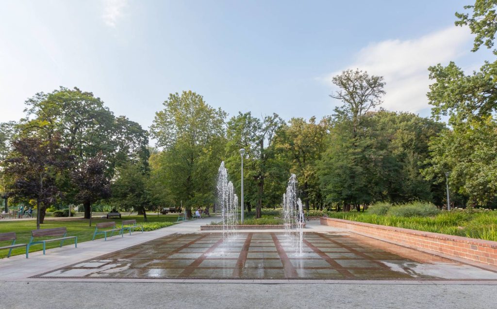 Park Lesława Węgrzynowskiego- Widok na fontannę, po lewej stronie cztery ławki z oparciem, po prawej stronie roślinność w ceglanych donicach. W tle zadrzewienie.