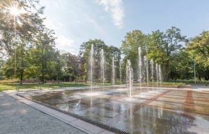 Park Lesława Węgrzynowskiego- zbliżone ujęcie na fontannę, na drugim planie dwie ławki i towarzyszące zadrzewienie.
